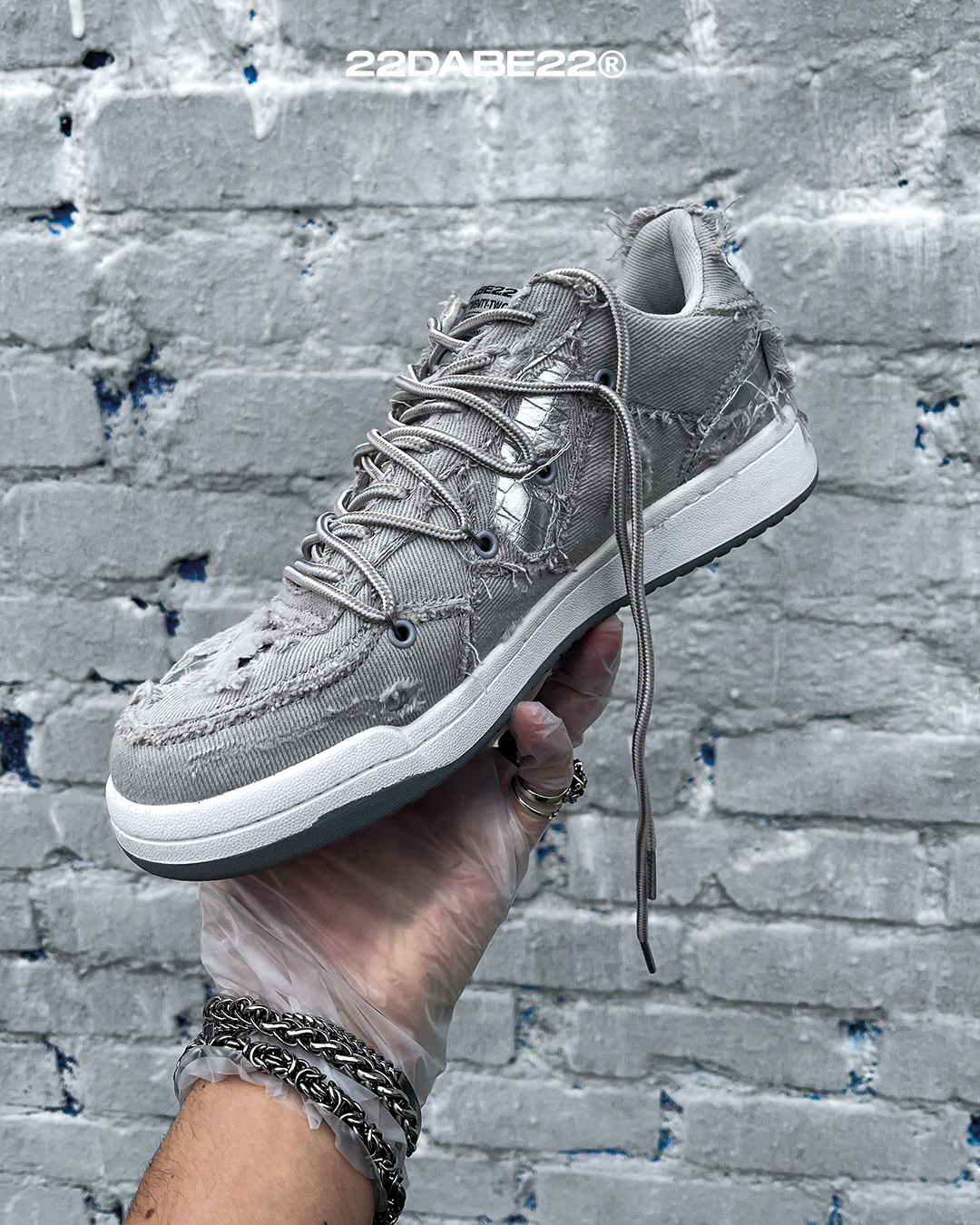 The DABE RANGER TWENTYTWO GREY shoe, a streetwear statement piece with metallic details under denim fabric on the upper. The shoe features double laces for extra style and versatility. Each shoe is handmade and unique, with every hole made by hand, giving it a one-of-a-kind look. The shoe is perfect for those who want to stand out from the crowd and make a fashion statement.